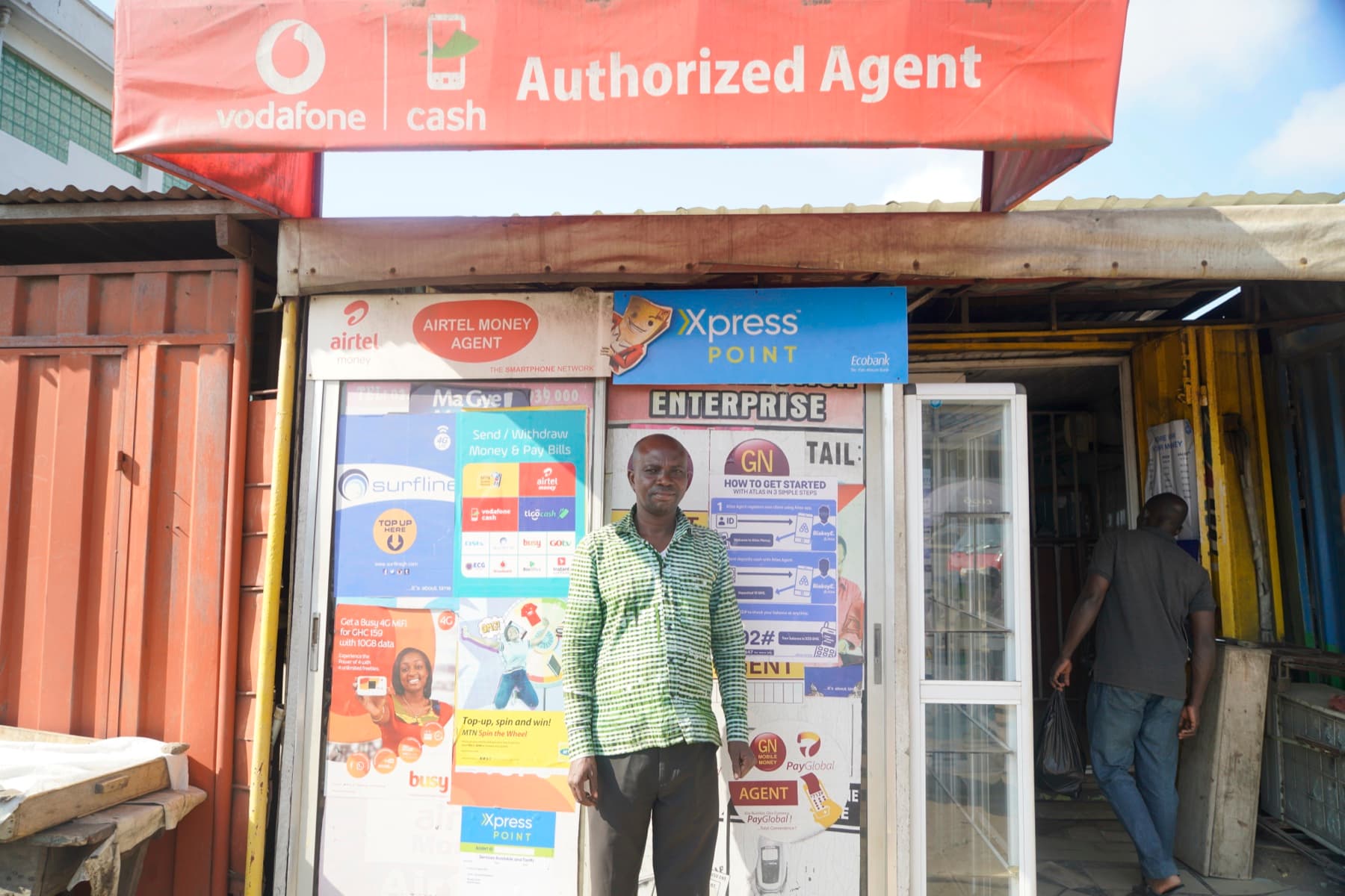 A mobile-money agent in front of his shop in Ghana, under Vodafone Cash and Airtel Money signs, with an Atlas 'how to get started in 3 simple steps' poster beside him.