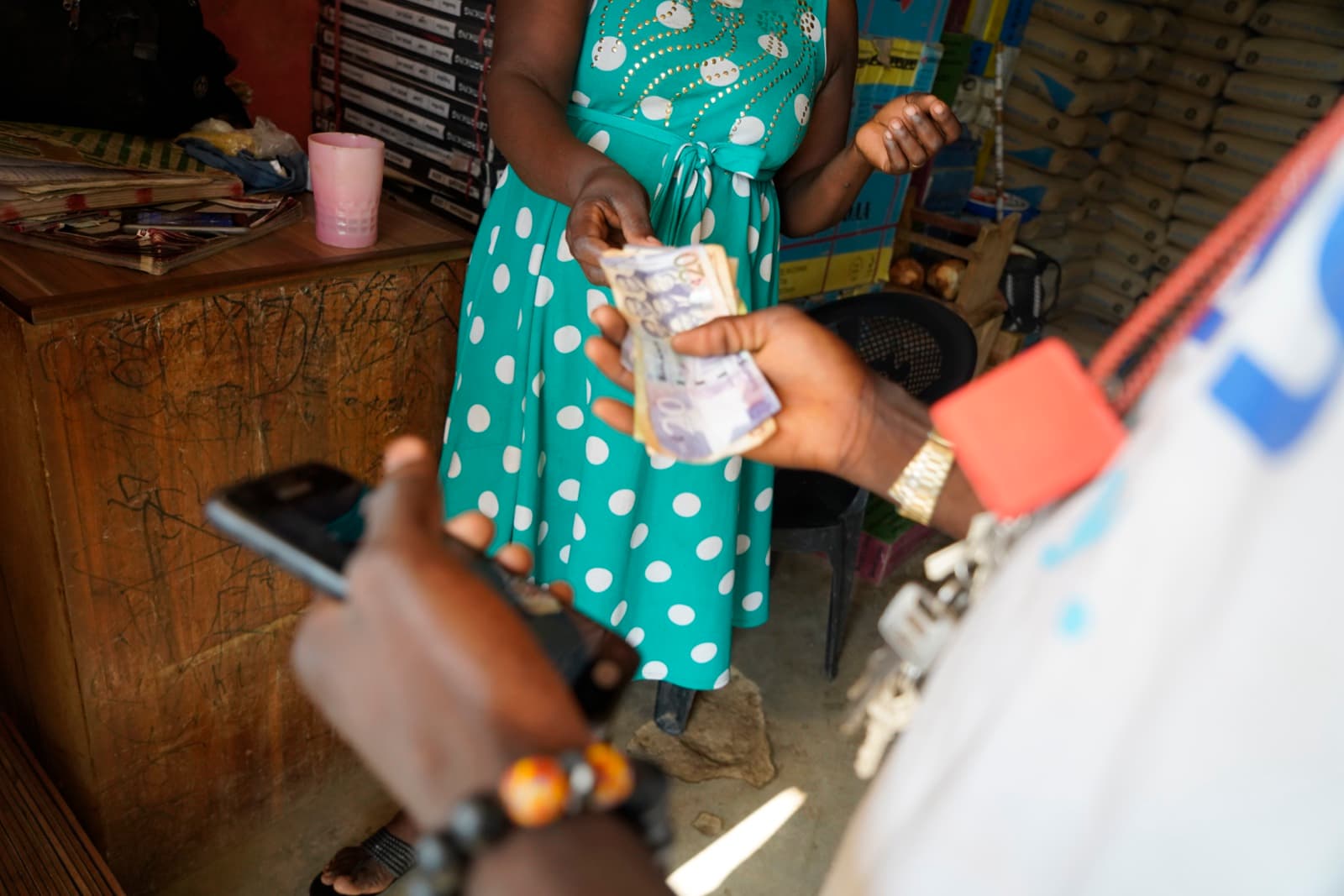 Cedi notes changing hands over a shop counter while the customer holds a feature phone.