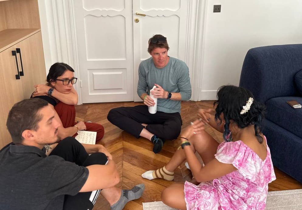Four people sitting cross-legged on the parquet floor of the apartment, one with a notebook, in conversation.